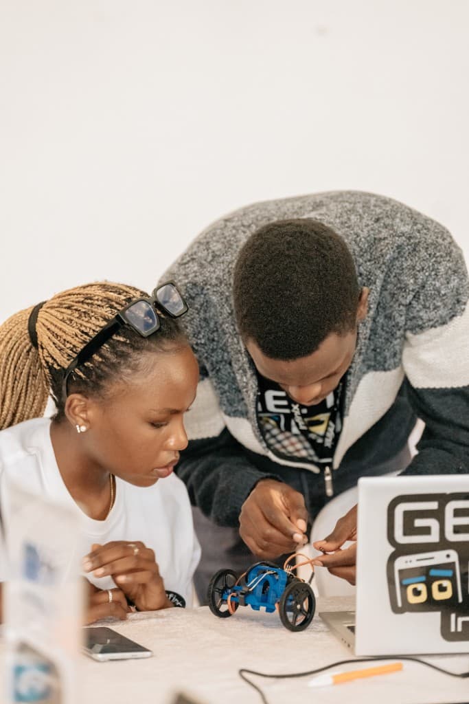 Two people collaborating at a table while assembling a small wheeled robot prototype